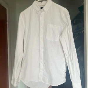 Vit linneskjorta - Säljer en stilren vit linneskjorta med klassisk krage och bröstficka. Skjortan har långa ärmar och knappar framtill. Perfekt för en avslappnad men ändå uppklädd look.