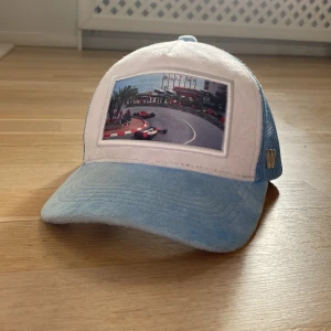 Blå och vit keps från Wonti - Snygg keps från Wonti i blått och vitt med ett coolt racingmotiv på framsidan. Kepsen har ett nätmaterial på sidorna och en böjd skärm. Perfekt för dig som gillar motorsport!