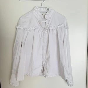Vit blus från Levate  - Säljer en stilren vit blus från Levete Room med volangdetaljer vid krage och ärmslut. Blusen har långa ärmar och knappar framtill. Perfekt för en elegant look.
