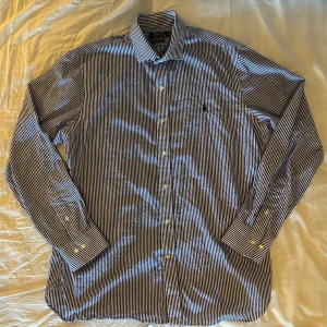 Randig skjorta från Ralph Lauren - Snygg randig skjorta från Ralph Lauren i blått och vitt. Skjortan har en slim fit passform och är tillverkad i 100% bomull med stretch. Den har långa ärmar och knappar framtill samt en broderad logga på bröstet. Storlek 17/43 vilket jag uppskattar motsvarar L. Inga fläckar. Knappt använd. 