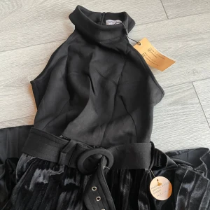 Svart ärmlös klänning med bälte stl 34 - Elegant svart ärmlös klänning med hög krage och ett matchande bälte i midjan. Kjolen är plisserad och ger en fin rörelse. Perfekt för en stilren look.