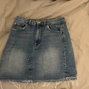 Jeanskjol från Gina  - Snygg jeanskjol från gina tricot i klassisk blå denim. Kjolen har en rå kant nertill och femficksdesign med knapp och dragkedja framtill. Perfekt för en avslappnad stil.