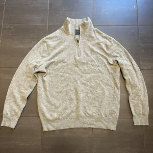 Ralph lauren - Säljer en stilren grå tröja från Ralph Lauren med halv dragkedja och bruna armbågslappar. Perfekt för kyligare dagar med sin mjuka och bekväma passform. Tröjan har långa ärmar och ribbade muddar.