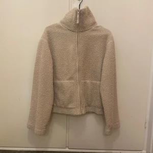 Beige teddyjacka med dragkedja - Mysig beige teddyjacka med dragkedja framtill. Jackan har en hög krage och långa ärmar, perfekt för kyliga dagar. Den fluffiga texturen ger en trendig look och den neutrala färgen gör den lätt att matcha med olika outfits.