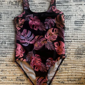Svart baddräkt med bladmönster - Snygg svart baddräkt med tropiskt bladmönster i rosa och lila nyanser. Baddräkten har breda axelband och en klassisk skärning. Setet är komplett med ett bad överdrag i ett väldigt lätt tyg