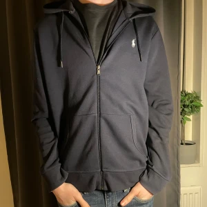 Mörkblå hoodie från Ralph Lauren - Säljer en stilren mörkblå hoodie från Ralph Lauren med dragkedja och huva. Tröjan har långa ärmar och en liten logga på bröstet. Perfekt för en avslappnad look. Storlek M