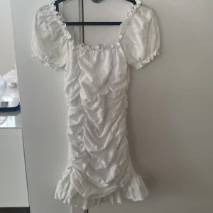 Vit klänning från Gina Tricot - Charmig vit klänning från Gina Tricot med puffiga kortärmar och volangdetaljer. Klänningen har ett vackert broderat mönster och en rynkad design som ger en romantisk känsla. Perfekt för sommarens festligheter!
