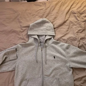 Grå hoodie från Ralph Lauren - Säljer en stilren grå hoodie från Ralph Lauren med dragkedja och klassisk logga på bröstet. Perfekt för en avslappnad look. Den har en praktisk huva med dragsko och fickor framtill.