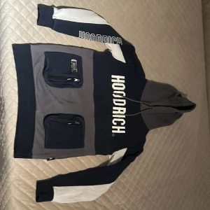 Blå och grå hoodie från Hoodrich - Säljer en snygg blå och grå hoodie från Hoodrich med vit text på bröstet. Den har en stor ficka framtill med två dragkedjor och en justerbar huva. Perfekt för en avslappnad stil.