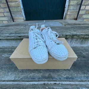 Vita sneakers med nitar från Christian Louboutin - Säljer ett par vita sneakers från Christian Louboutin med coola nitar på tån och den ikoniska röda sulan. Skorna har snörning och är perfekta för att ge din stil en edgy touch. Kommer med originalkartong.