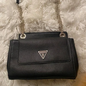 Svart axelväska från Guess - Snygg svart axelväska från Guess med kedjedetaljer och logotyp på framsidan. Väskan har en praktisk storlek och stängs med ett vridlås. Perfekt för att bära dina nödvändigheter med stil.