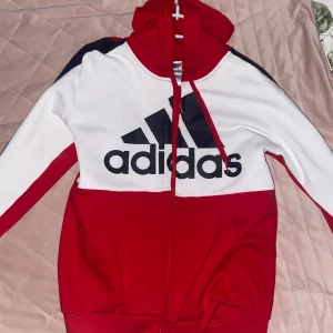 Röd och vit hoodie från Adidas - Snygg röd och vit hoodie från Adidas med dragkedja och stor logga på framsidan. Tröjan har långa ärmar och en bekväm huva med snören. Perfekt för en sportig look!