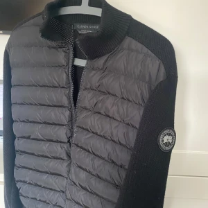 Svart Cardigan från Canada Goose - Säljer en sällsyns svart Canada Goose black label cardigan med quiltad framsida och stickade ärmar. Jackan har en dragkedja framtill och en rund krage. Perfekt för kyligare vårdagar eller sena kvällar. Jackan är helt oanvänd, Hör av er vid intresse!