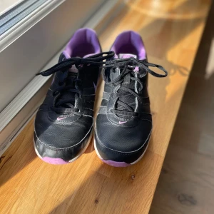 Svarta och lila Nike sneakers - Snygga svarta Nike air sneakers med lila detaljer och vit sula. Skorna har snörning och en bekväm passform, perfekt för vardagsbruk eller ett träningspass. Den lila insidan och Nike-loggan ger en cool kontrast till det svarta.