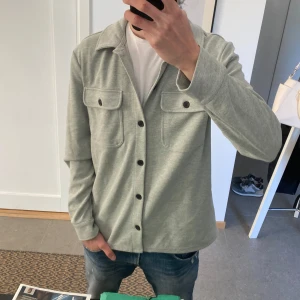 Grå overshirt med knappar - Snygg grå overshirt med långa ärmar och knappar framtill. Två bröstfickor med knappar ger en stilren look. Perfekt för lager-på-lager-stil.