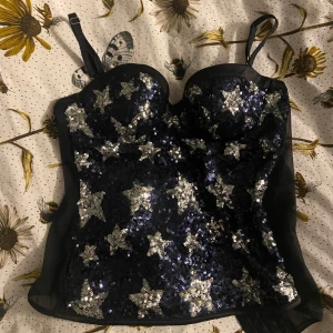 Svart paljettkorsett med stjärnor - Snygg svart korsett topp täckt med glittriga stjärnformade paljetter. Perfekt för en festlig kväll. Den har justerbara axelband och en figurnära passform.