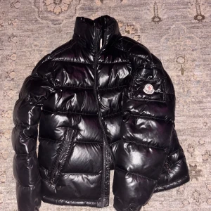 Moncler Maya - Säljer en snygg svart dunjacka från Moncler. Jackan har en glansig finish och är försedd med en dragkedja framtill. Den har långa ärmar och en praktisk ficka med Moncler-logga på ärmen. Perfekt för kyligare dagar. Jackan har dessvärre ingen luva. Vilket ger den skicket 7/10. Priset är hugget i sten. Skriv vid funderingar!