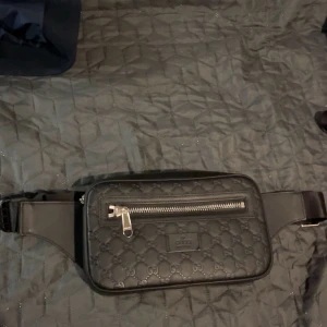 Svart Gucci midjeväska med mönster - Snygg svart midjeväska med präglat mönster och dragkedja framtill. Väskan har justerbart bälte och är perfekt för att hålla dina saker säkra och nära till hands. Den har en stilren design med en praktisk ficka på framsidan.     ❗️❗️❗️❗️ Pris är diskutabelt vid en snabb affär ❗️❗️❗️❗️