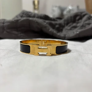 H armband - Stilrent H armband i guldpläterad metall med svarta emaljdetaljer. Armbandet har den ikoniska H-formade låset framtill. 