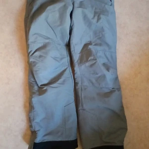 Grå täckbyxor från Wildriver - Säljer ett par grå täckbyxor från Wildriver med svarta detaljer vid bensluten. Byxorna har dragkedja och knäppning framtill samt justerbar midja med kardborre. Perfekta för kalla dagar och vinteraktiviteter.