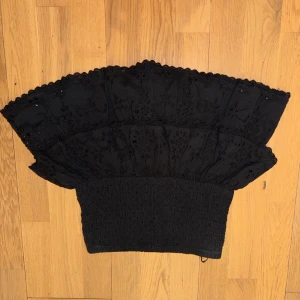 Svart volang tubtopp från Gina Tricot - Supersnygg svart tubtopp från Gina Tricot med bred smockad midja och två lager volanger upptill. Toppens tyg har broderade detaljer och spetskant som ger en extra cool vibe. Perfekt att styla med jeans eller kjol för en trendig look.