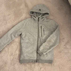Sail Racing ZIP hoodie  - Snygg ljusgrå zip hoodie från Sail Racing med dragkedja framtill och på bröstfickan. Broderad logga på bröstet och huvan. Mjuk insida och ribbade muddar. Perfekt för chill dagar eller när du vill ha en sportig look. Väldigt bra skick då den är använd väldigt få gånger (ca 2-3)!!! 