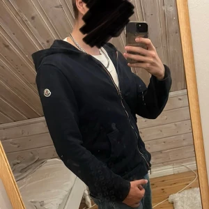 Mörkblå hoodie från Moncler - Säljer en stilren mörkblå hoodie från Moncler med huva och dragkedja framtill. Tröjan har långa ärmar, två fickor och Moncler-logga på vänster ärm. Materialet är mjuk bomull som känns skönt mot huden. Perfekt för dig som gillar klassisk streetwear med premiumkänsla.