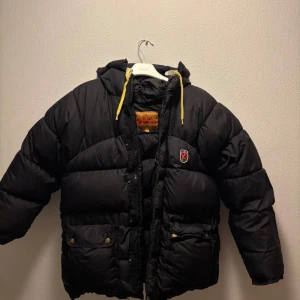 Svart dunjacka från Fjällräven - Säljer en svart dunjacka från Fjällräven med huva och gula snören. Jackan har en broderad logotyp på bröstet och praktiska fickor med knappar. Perfekt för kalla vinter dagar. En liten bit där den är ihopsydd vilket inte syns.Storlek XXS. PRIS KAN DISKUTERAS! Kan även byta mot en annan Fjällräven jacka 