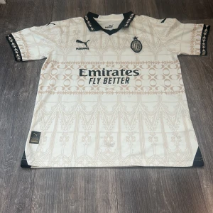 AC Milan vit kortärmad fotbollströja Puma - AC Milan fotbollströja från Puma i vit med ljusbeige grafiskt mönster och svarta detaljer på krage och ärmslut. Tryckta loggor för Emirates och Pleasures på framsidan, samt wefox på baksidan. Tröjan har en klassisk passform och är tillverkad i lätt polyester.