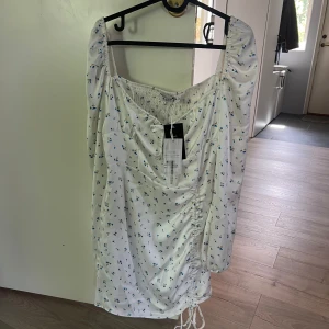 Vit blommig klänning från NA-KD - Supersöt vit kortklänning från NA-KD med små blå blommor, långa puffärmar och smock i ryggen. Klänningen har snörning framtill och rynkade detaljer längst ner. Prislappen sitter kvar och klänningen är alltså aldrig använd, säljer den endast för att den är för liten. Normal i storleken 