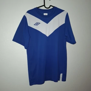 Blå och vit Umbro fotbollströja S - Snygg blå fotbollströja från Umbro med vitt V-format fält över bröstet och klassisk logga. Tröjan är kortärmad och gjord i ett lätt och ventilerande material som passar perfekt till träning eller match.