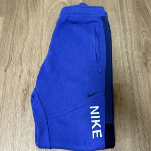 Blåa mjukisbyxor från Nike - Snygga blå mjukisbyxor från Nike med dragkedjeficka och logga på benet. Byxorna har resår i midjan och är perfekta för chill eller träning. Materialet är mjukt och skönt, och färgen är riktigt poppig blå.