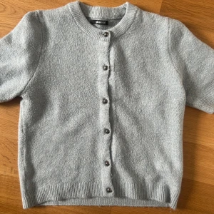 Ljusblå kofta från Gina Tricot - Kortärmad ljusblå kofta från Gina Tricot med rund halsringning och knappar framtill. Koftan är stickad och har ribbade muddar vid hals och nederkant. Perfekt att slänga över en topp för en chill look.