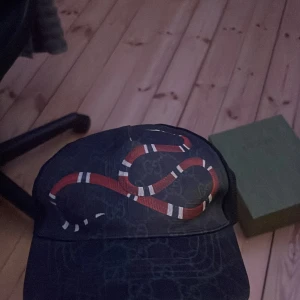 Gucci keps med orm och GG-mönster - Snygg svart keps från Gucci med klassiskt GG-mönster och en röd-vit orm broderad framtill. Justerbar rem baktill med röd och marinblå rand. Kepsen är tillverkad i bomull och mesh för en luftig känsla. Kommer med originalbox, dustbag och tags. Pris kan diskuteras!