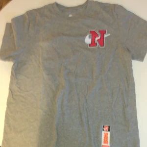 Grå Nike t-shirt med tryck - Grå t-shirt från Nike med coola tryck i rött, vitt och marinblått både fram och bak. Stor röd N med vit swoosh på bröstet och flera grafiska Nike-motiv på ryggen. Rund hals och korta ärmar. Materialet känns som mjuk bomull, perfekt för en chill streetwear-look.