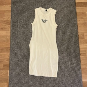 Vit ärmlös klänning från DIVIDED - Säljer en vit, tight och ärmlös kortklänning från DIVIDED. Klänningen har en rund hals och ett svart tryck framtill vid bröstet. Materialet är mjukt och stretchigt, perfekt för dig som gillar minimalistisk stil med en edgy touch.