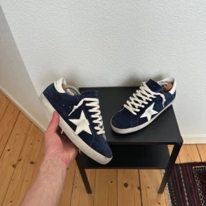 Blå mocka sneakers med vit stjärna - Säljer ett par Golden Goose Superstar sneakers i mörkblå mocka med vit stjärndetalj på sidan och vita snören. Skorna har rund tå, platt sula och cool retrovibe. Insidan är märkt med 'SUPERSTAR' och de är tillverkade i Italien. Perfekt för dig som gillar unik streetstil.