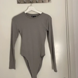 Grå långärmad bodysuit från Zara - Säljer en stilren grå långärmad bodysuit från Zara. Den har rund halsringning och är tillverkad i mjuk bomullsmix som sitter snyggt mot kroppen. Perfekt att styla med jeans eller kjol för en clean look. (Aldrig använd)