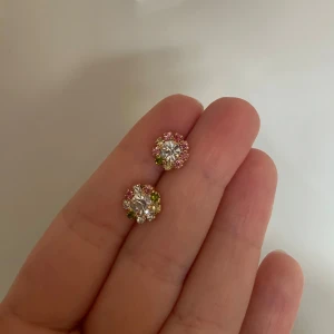Färgglada glittriga örhängen - Säljer ett par runda örhängen med stor klar sten i mitten och små rosa, gröna och gula stenar runt om. Örhängena har en glittrig look och guldtonad infattning. Perfekta för dig som gillar färg och bling!