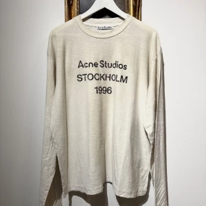 Vit långärmad tröja Acne Studios - Långärmad beige tröja från Acne Studios med svart tryck framtill där det står 'Acne Studios STOCKHOLM 1996'. Tillverkad i en mjuk blandning av bomull och hampa. Avslappnad passform och rund halsringning, perfekt för en clean och stilren look.