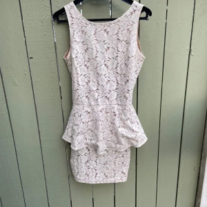 Vit spetsklänning med volang - Supersöt vit kortklänning med blommig spets och volang i midjan. Klänningen är ärmlös och har en snygg v-ringad rygg med dragkedja. Perfekt för sommarens alla tillfällen när du vill känna dig extra fin.34-38