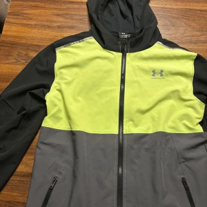 Under Armour vindjacka med huva - Sportig vindjacka från Under Armour i svart, grått och limegrönt. Jackan har huva, hel dragkedja framtill och två sidofickor med dragkedja. Perfekt för utomhusaktiviteter och träning, med lätt och smidigt material som andas.14 år men lite liten i storlek Helt ny.