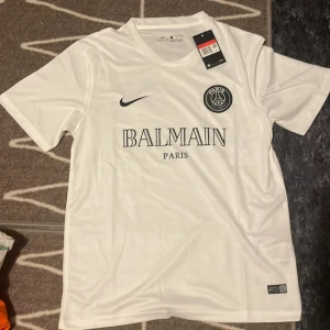 Vit PSG x Balmain fotbollströja Nike - Snygg vit fotbollströja från Nike i samarbete med PSG och Balmain. Tröjan har korta ärmar, rund hals och tryckta loggor på bröstet. Tillverkad i lätt och ventilerande material som passar perfekt för träning eller match.