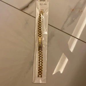Guldigt armband i rostfritt stål - Snyggt guldigt armband med länkdesign i rostfritt stål. Armbandet har en rektangulär platta med gravyr och en klassisk, chunky kedja som ger en cool och modern vibe. Perfekt för dig som gillar statement-accessoarer och vill sticka ut.