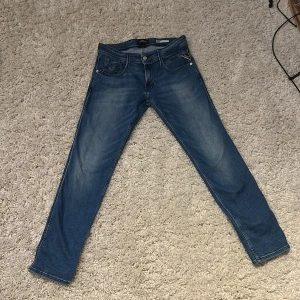 Blå Replay Anbass jeans herr - Snygga blå Replay Anbass jeans med klassisk femficksdesign och diskret slitning på låren. Jeansen har en slim passform och raka ben, tillverkade i mjukt bomullstyg med lite stretch för extra komfort. Perfekta för dig som gillar stilrena och moderna jeans.