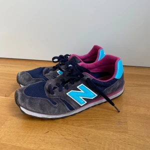 New Balance sneakers i blå och grå - Snygga New Balance sneakers med mörkblå mesh, grå mockadetaljer och en stor turkos N-logga på sidan. Insidan är fodrad i cerise och sulan har en vit och rosa kant. Perfekta för dig som gillar färg och sportig stil. Köpta på second hand så hade en sliten look från början!