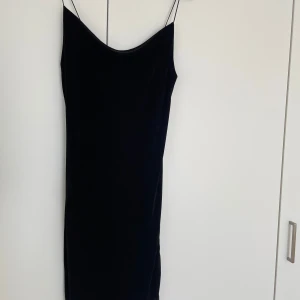 Svart långklänning med tunna axelband - Snygg svart långklänning med tunna axelband och rak siluett. Klänningen har en enkel och stilren design med lätt glansigt tyg som ger en lyxig känsla. Perfekt för dig som gillar minimalistisk stil och vill ha en klänning som sticker ut på ett subtilt sätt.