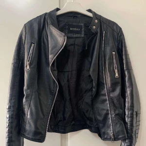 Svart skinnjacka från Missay - Snygg svart skinnjacka från Missay med coola dragkedjedetaljer och quiltade partier på axlar och ärmar. Jackan har en klassisk bikerstil med ståkrage och flera fickor med dragkedja. Perfekt för dig som vill ha en edgy look.
