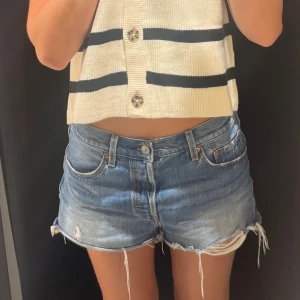 Blå jeansshorts med fransad kant - Säljer ett par klassiska blå jeansshorts med fransad och lätt sliten kant. Modellen är midwaist med fem fickor och knappgylf. Perfekta för chill dagar och har en loose passform som ger en avslappnad vibe.