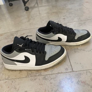 Nike Air Jordan 1 Low svart/grå/vit - Snygga Nike Air Jordan 1 Low i svart, grått och vitt skinn. Klassisk siluett med svart swoosh, perforerad tåbox och broderad Air Jordan-logga på hälen. Låga sneakers med svart snörning och vit sula – perfekt för dig som gillar streetstyle.